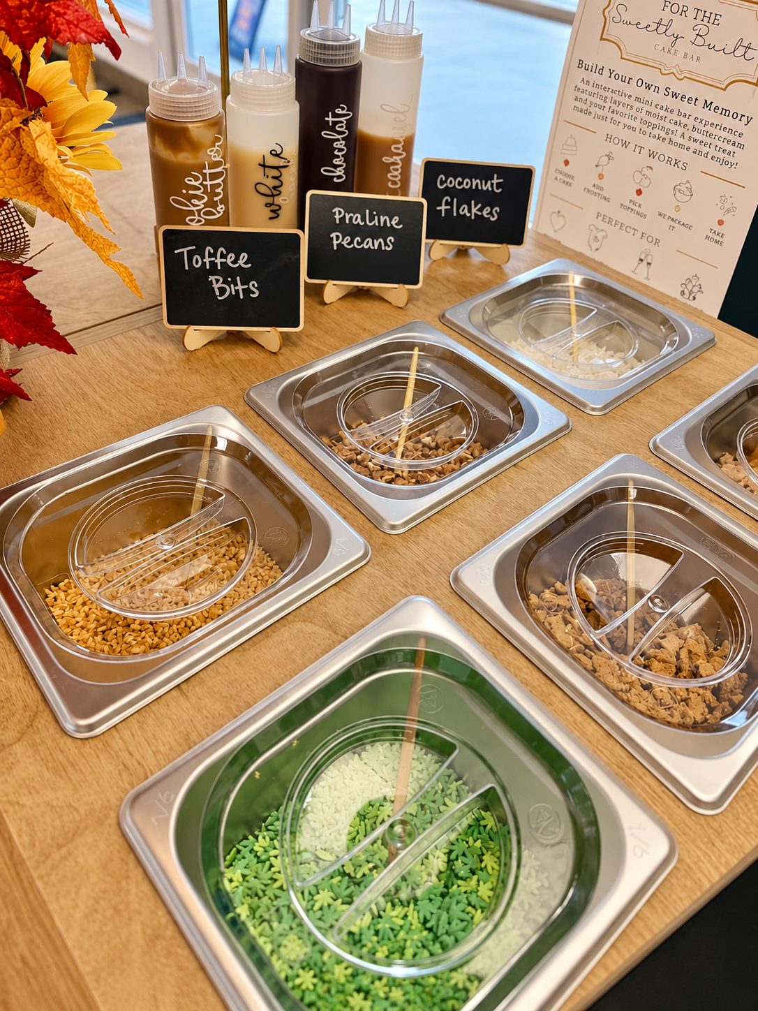 Sweetly Built cake bar topping wells filled with leaf sprinkles, toffee bits, praline pecans, coconut flakes, and crushed cookies, with drizzle bottles and the How It Works menu card behind