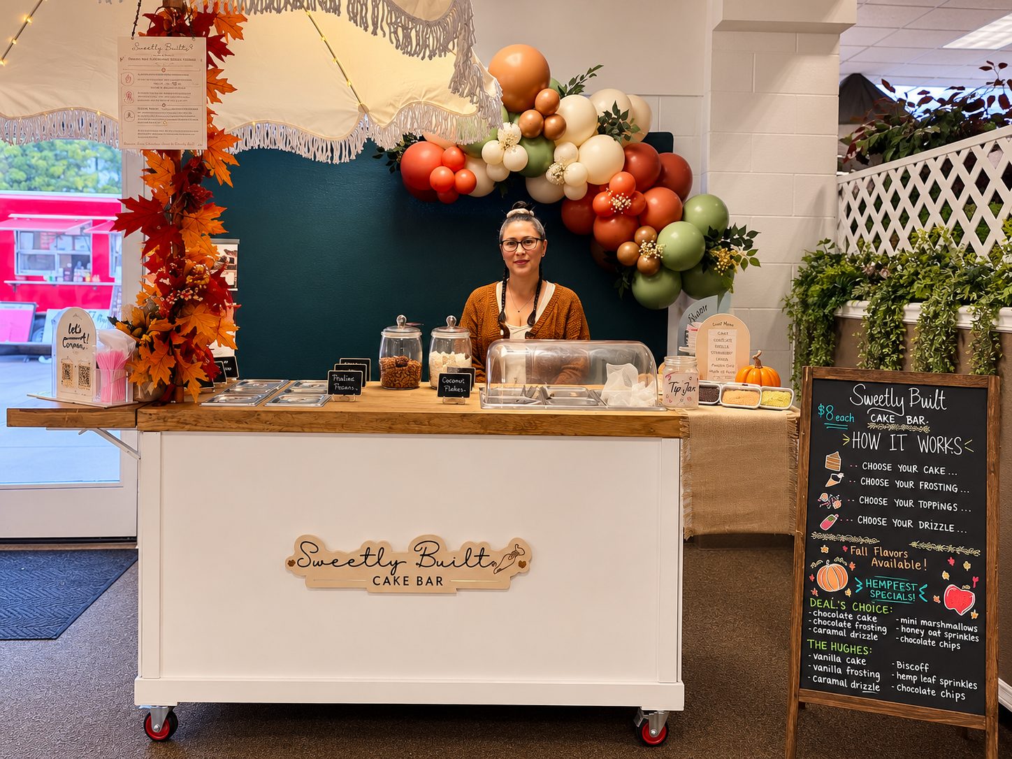 The Sweetly Built cake bar cart with fringed umbrella, balloon garland, and chalkboard menu set up against a teal wall at Treasure Valley HempFest